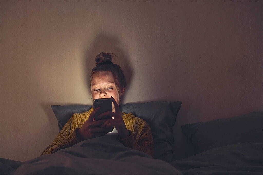 Mädchen liegt im Dunkeln in ihrem Bett und schaut auf ihr Smartphone-Display.